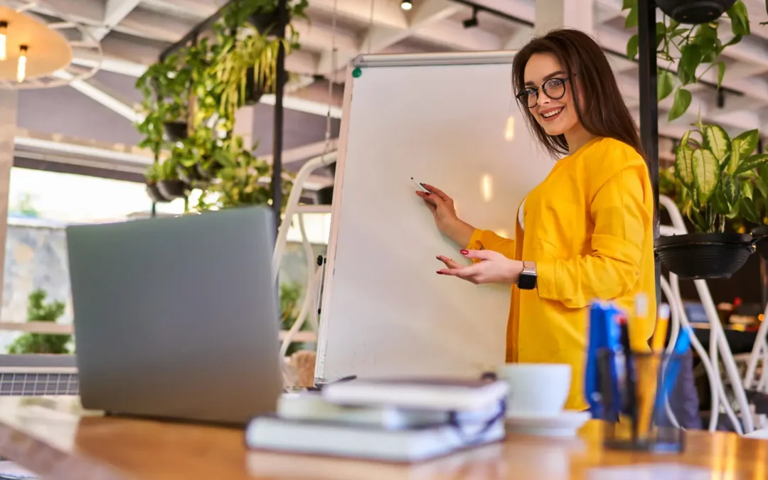 A woman in yellow is giving a presentation using a whiteboard in a modern, green workspace.